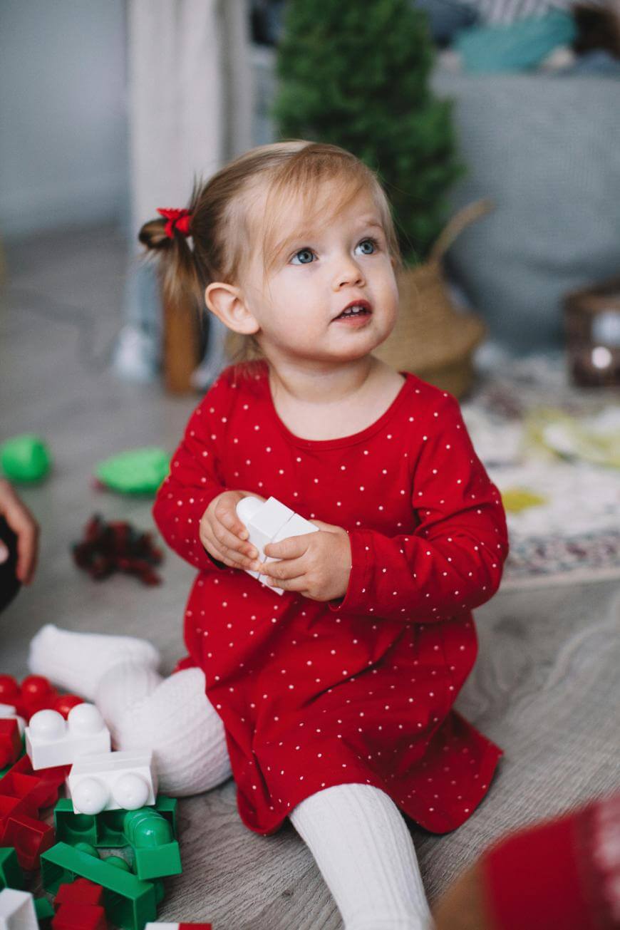 A young girl in a red dress with white polka dots sits on the floor, holding white toy blocks. She has light brown hair in pigtails and is surrounded by colorful building blocks.