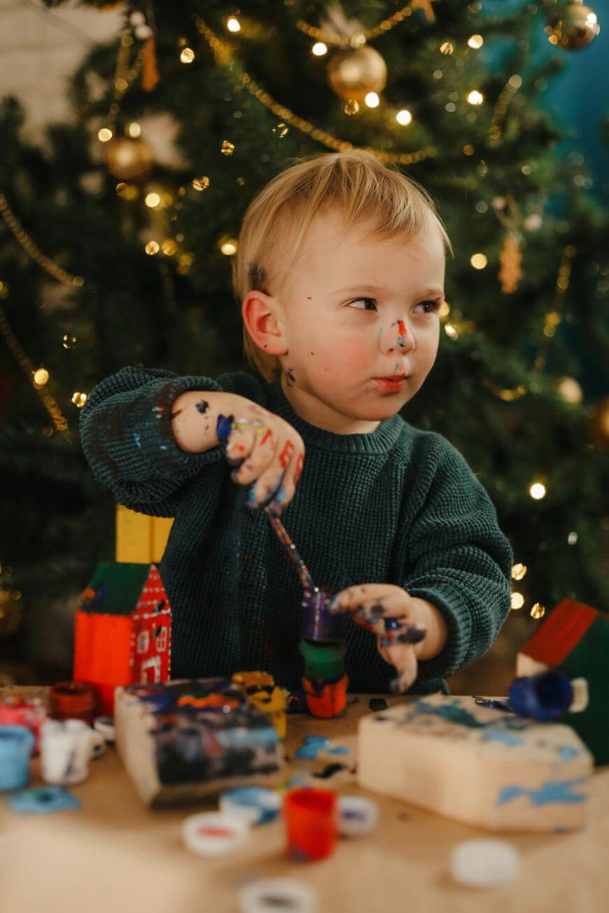 A young child with paint on their hands and face is painting wooden toys at a table. Behind them is a decorated Christmas tree with lights and ornaments. The scene looks festive and creative.