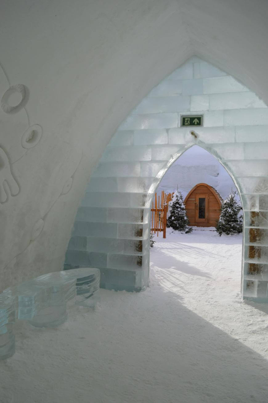 View from inside an ice building with arched doorway made of ice blocks, showing snow outside, small trees, and a wooden cabin in the background. There is a green exit sign above the doorway.