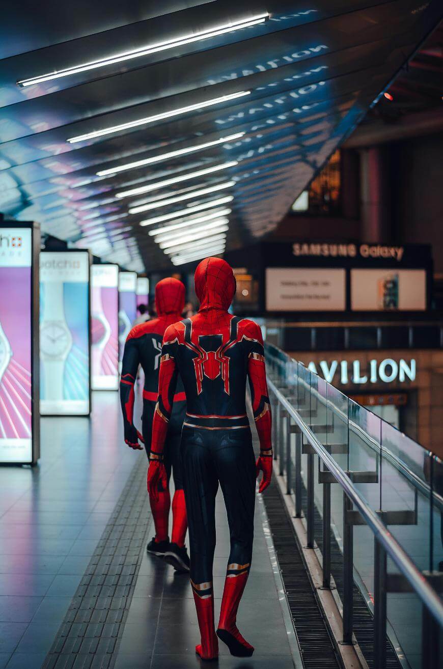 Two people dressed in Spider-Man costumes walk down a brightly lit, modern indoor walkway with illuminated signs and advertisements lining the passage.