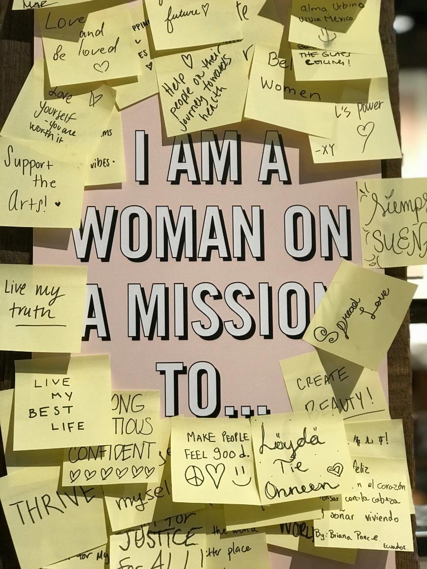 A sign reading I am a woman on a mission to... is surrounded by yellow sticky notes with handwritten empowering messages, goals, and affirmations about positivity, self-expression, and making a difference.