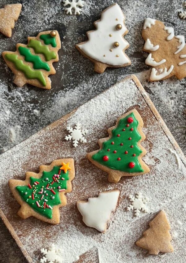 Christmas tree-shaped cookies decorated with green, red, and white icing and sprinkles rest on a wooden board dusted with powdered sugar, surrounded by small snowflake-shaped cookies.