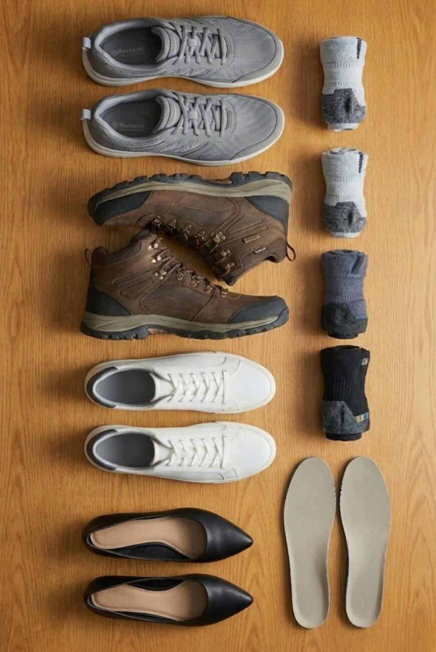 Four pairs of shoes—gray sneakers, brown hiking boots, white sneakers, and black flats—are neatly arranged on a wooden surface, accompanied by three pairs of rolled socks and one pair of beige insoles.