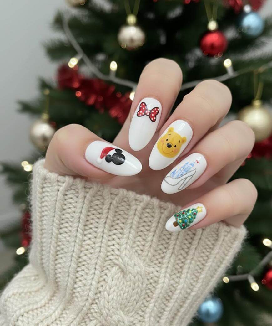 A hand with long, white manicured nails featuring various designs: Minnie Mouse bow, Mickey Mouse glove, Winnie the Pooh face, Cinderella’s glass slipper, and a decorated Christmas tree. A Christmas tree is in the blurred background.