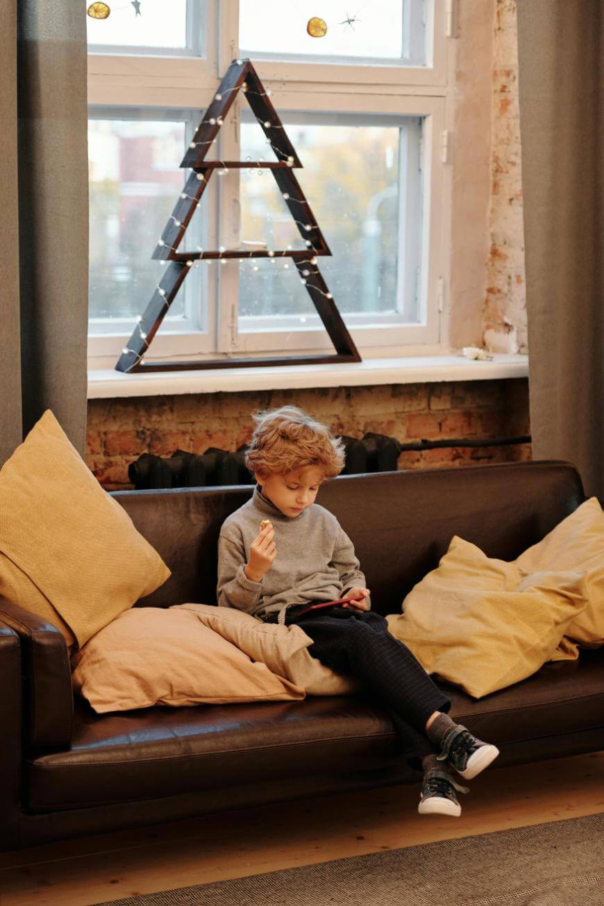 A young child with curly hair sits on a brown sofa with yellow pillows, looking at a tablet. Behind the child is a window with a triangular, tree-shaped decoration wrapped in lights.