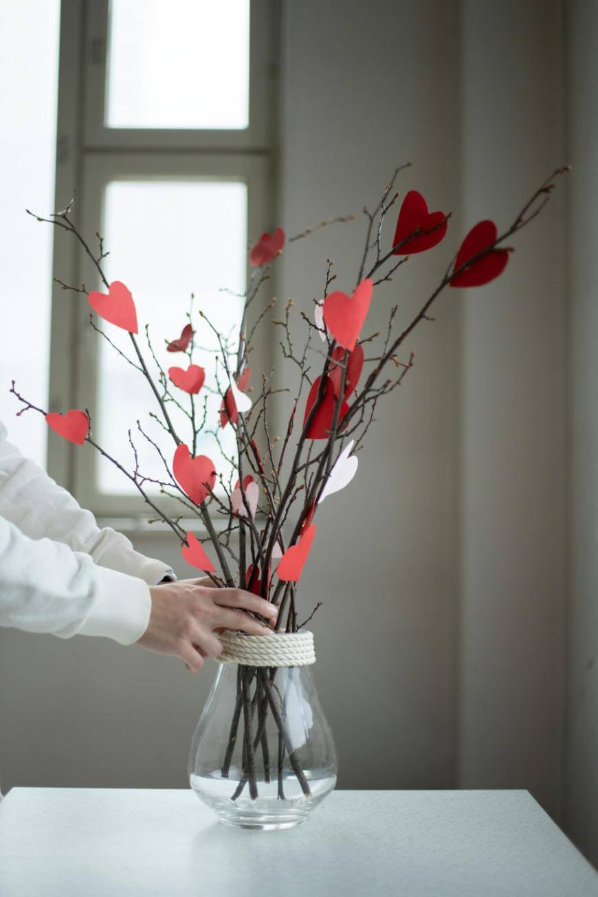 A person arranges bare branches with red and white paper hearts in a clear glass vase on a white table, with a window in the background.
