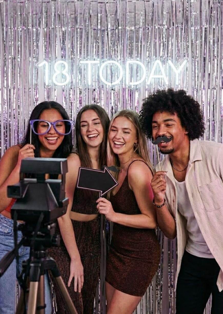 Four young adults pose and smile for a photo at a party, standing in front of shiny silver streamers and a neon sign that reads 18 TODAY. They hold fun props, including glasses, an arrow, and a mustache.