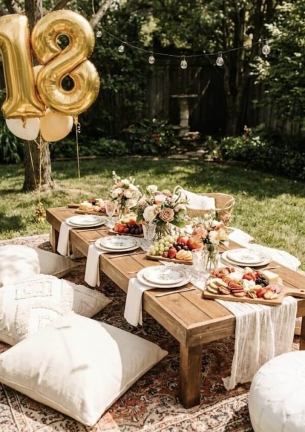 A low wooden table set outdoors with flowers, plates, and fruit trays, surrounded by floor cushions on a rug. Gold 18 balloons and string lights hang above, creating a festive, elegant picnic atmosphere.