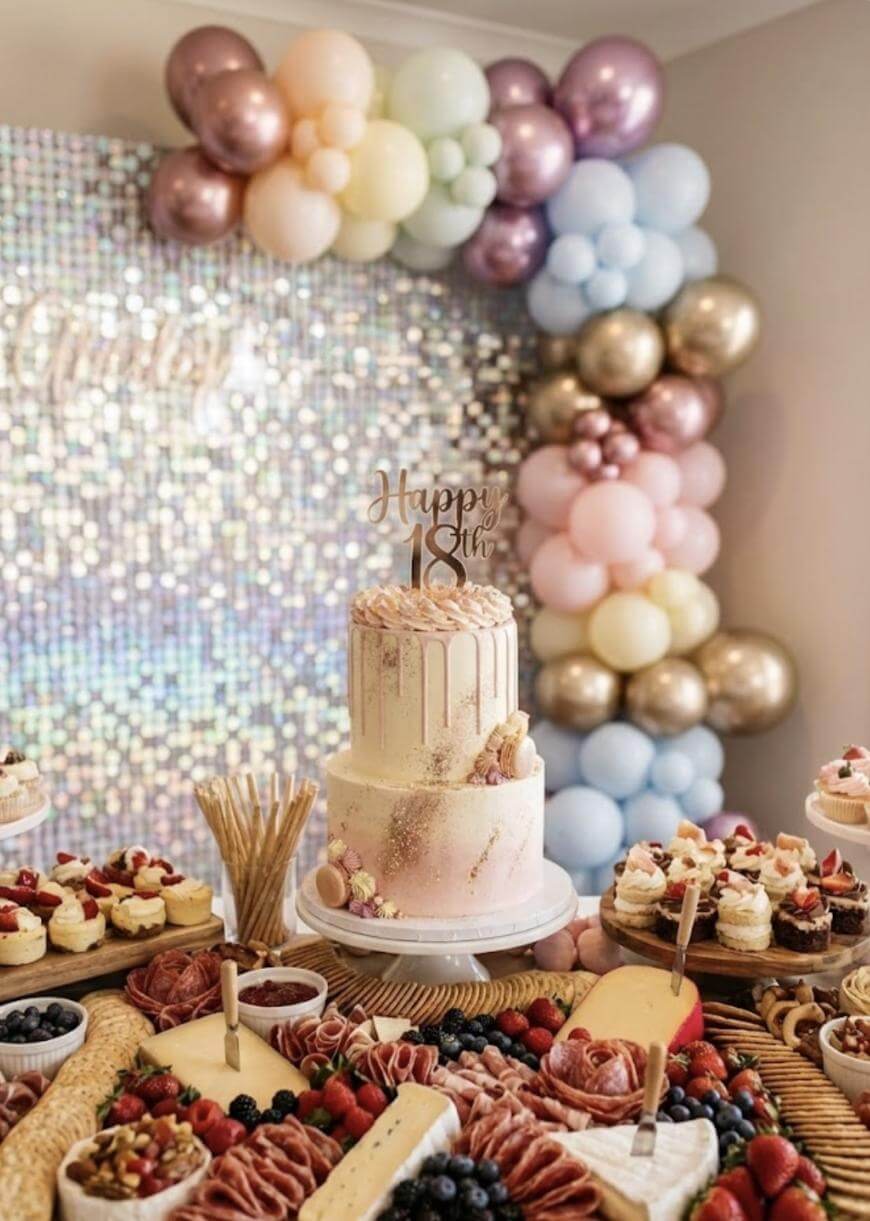 A decorated table with a pink “Happy 18th” cake, assorted pastries, fruit, cheese, and meats. Behind it, a shimmering backdrop and pastel balloon arch create a festive party setting.