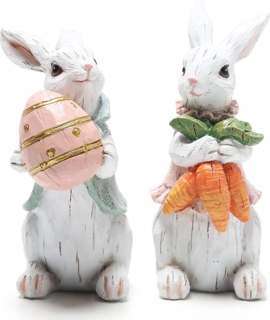 Two white bunny figurines; one holds a pink decorated egg, the other holds a bunch of orange carrots. Both have pastel-colored clothing and are posed upright.