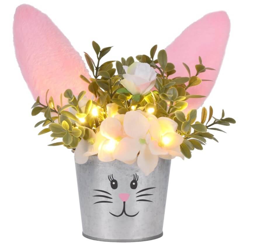 A metal flower pot decorated with a cute bunny face, pink furry ears, artificial flowers, green leaves, and small yellow LED string lights.