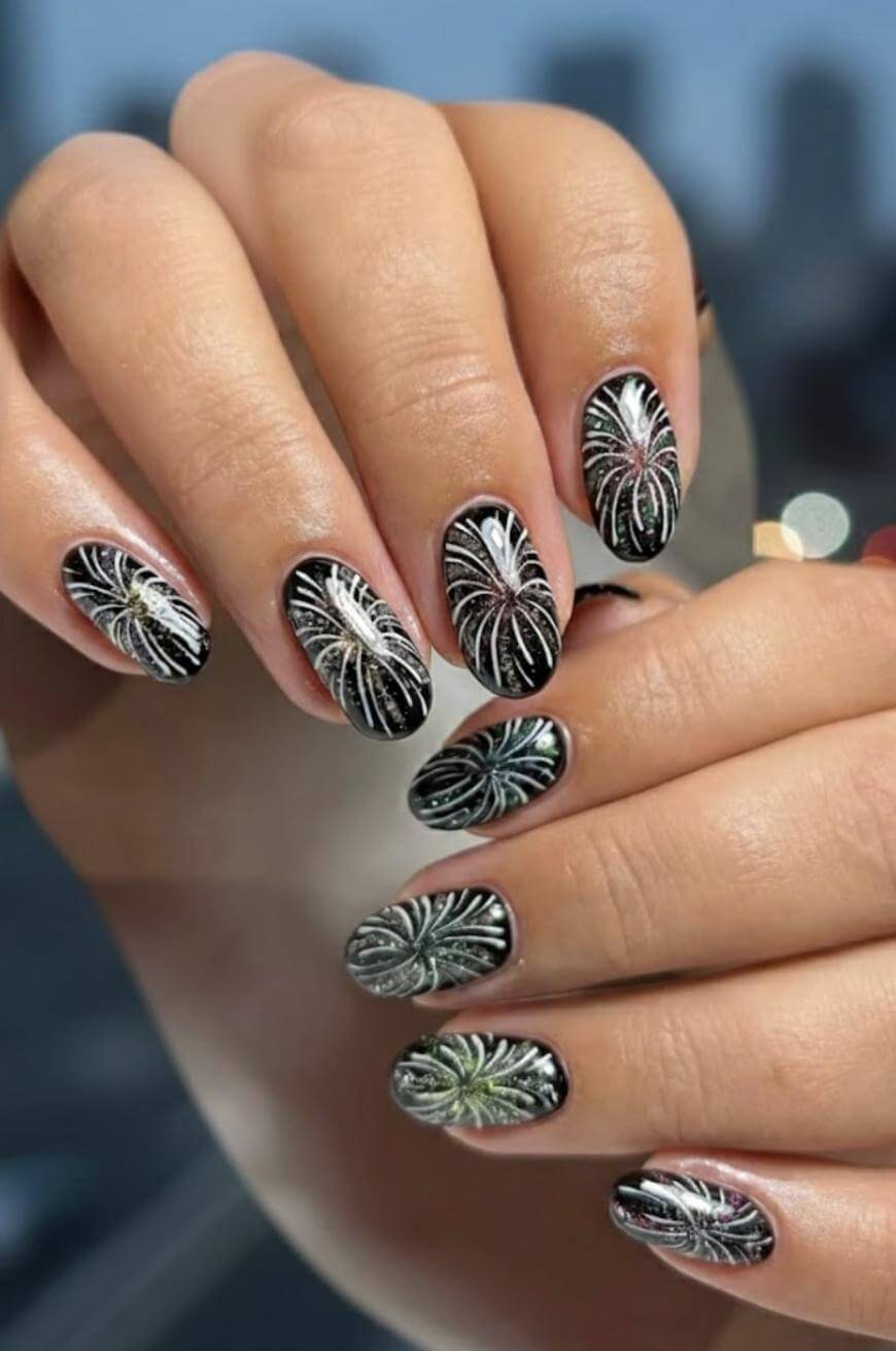 A hand with oval-shaped nails painted black, each nail decorated with silver and green firework-like designs. The background is blurred, highlighting the nail art.