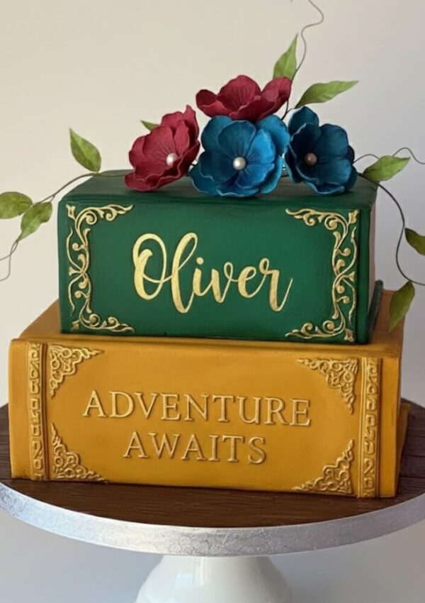 A two-tiered cake designed to look like stacked books: the top green book with Oliver in gold script and the bottom yellow book with ADVENTURE AWAITS in gold. Three red and blue flowers decorate the top.