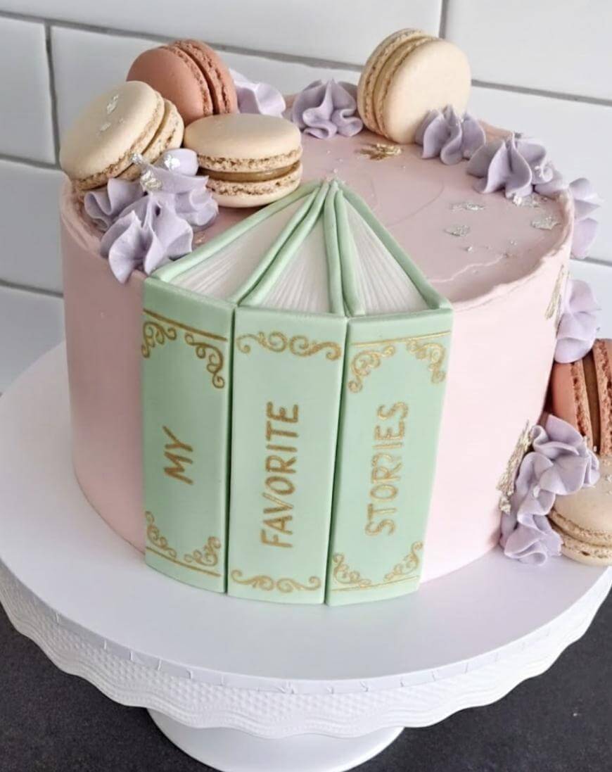 A pastel pink cake decorated with macarons and purple frosting swirls, featuring three edible books with green covers on the side that read My Favorite Stories in gold lettering. The cake sits on a white cake stand.