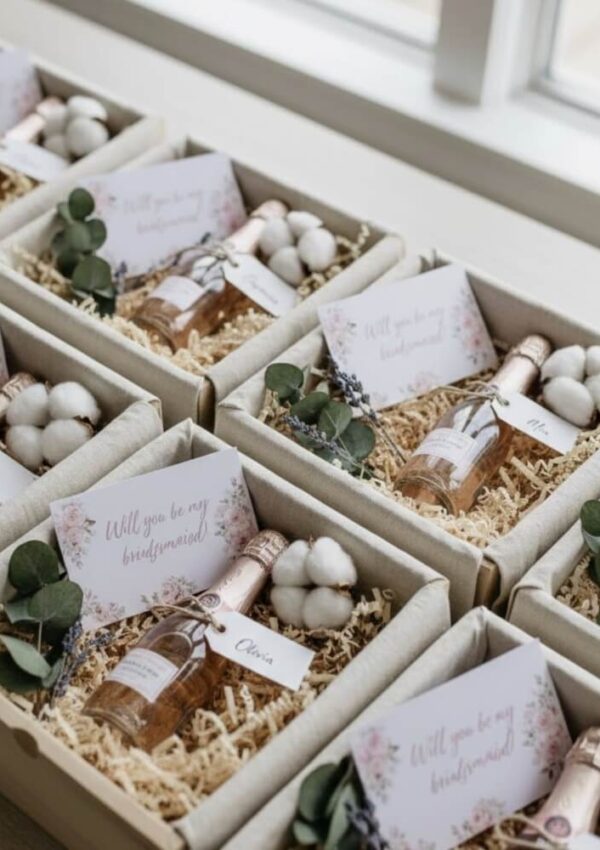 Several gift boxes with small bottles of rosé, sprigs of greenery, white decorative items, and cards reading Will you be my bridesmaid? are neatly arranged by a window.
