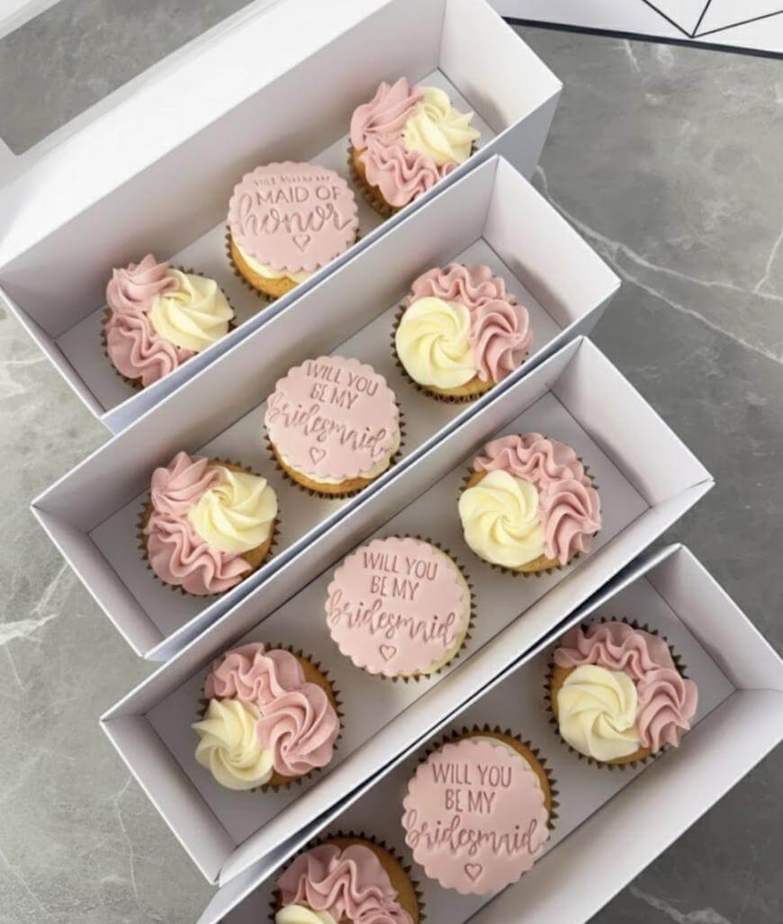Three white boxes, each containing four cupcakes with pink and cream swirled frosting. One cupcake per box has a round topper reading either Will you be my bridesmaid? or Maid of honor in elegant script.