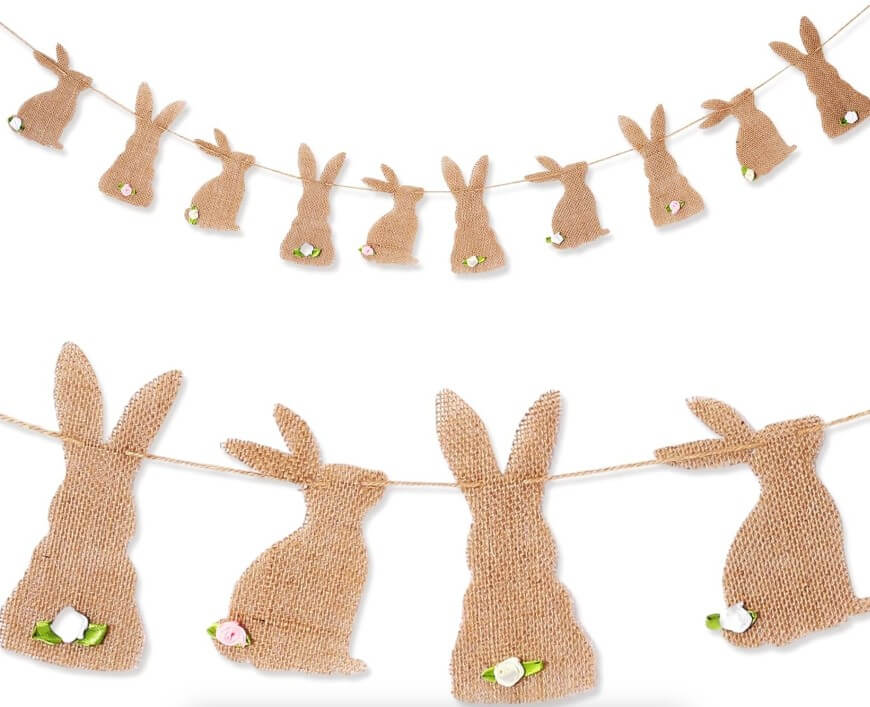 A garland featuring burlap bunny cutouts hanging from twine, each bunny adorned with a small flower decoration near its tail. The bunnies are arranged in a row, creating a festive, rustic decoration.
