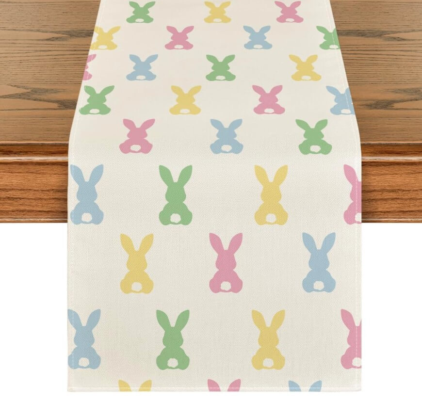 A white table runner decorated with pastel pink, blue, green, and yellow bunny silhouettes, each with a white tail, arranged in neat rows on a wooden table.