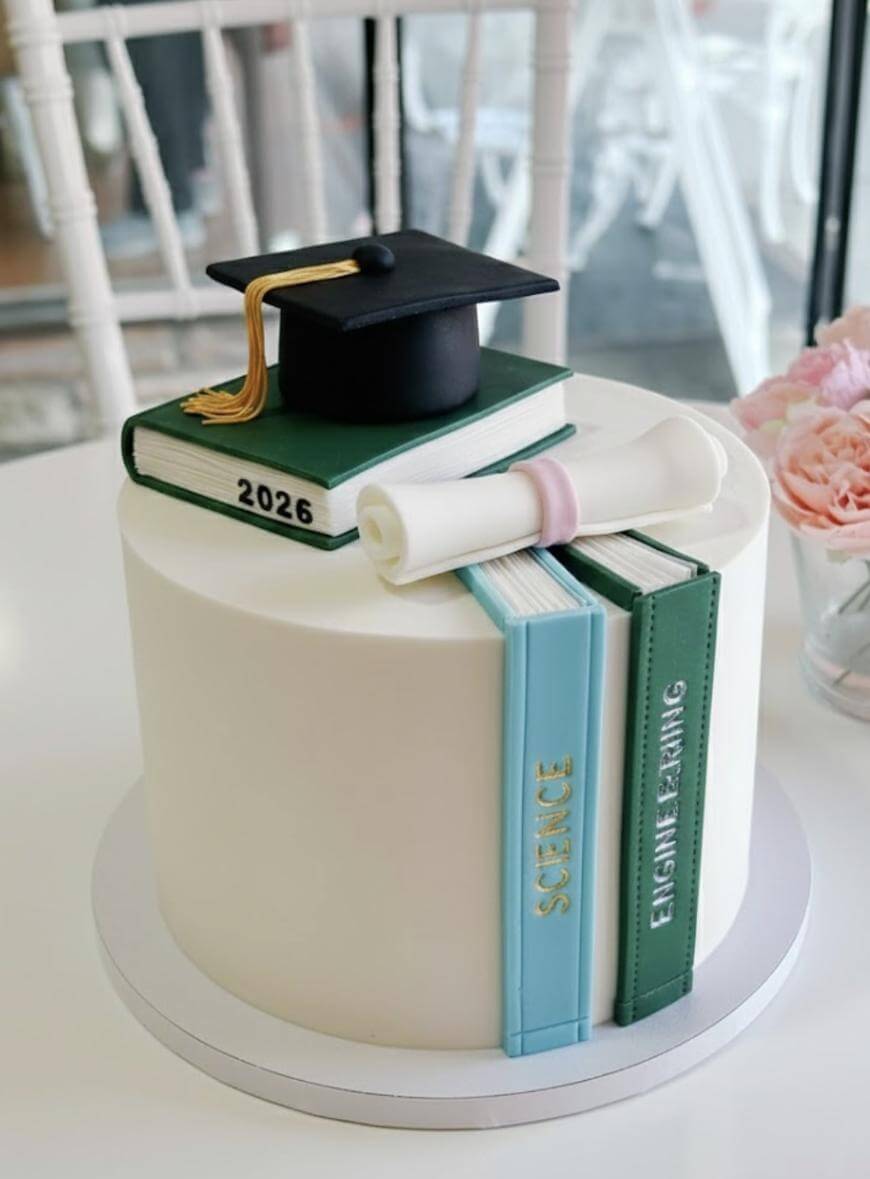 A round white cake decorated with edible books, a graduation cap, and a diploma. The books are labeled SCIENCE, ENGLISH LEARNING, and 2026. The cake sits on a white table near pink flowers.