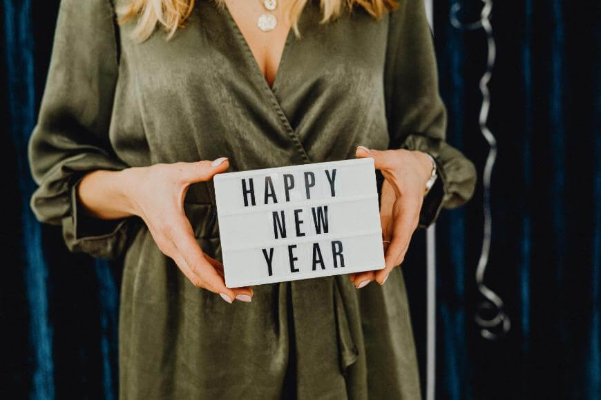 A person in a green outfit holds a white lightbox sign with the words HAPPY NEW YEAR in black letters. The background is dark, and festive decorations are partially visible.