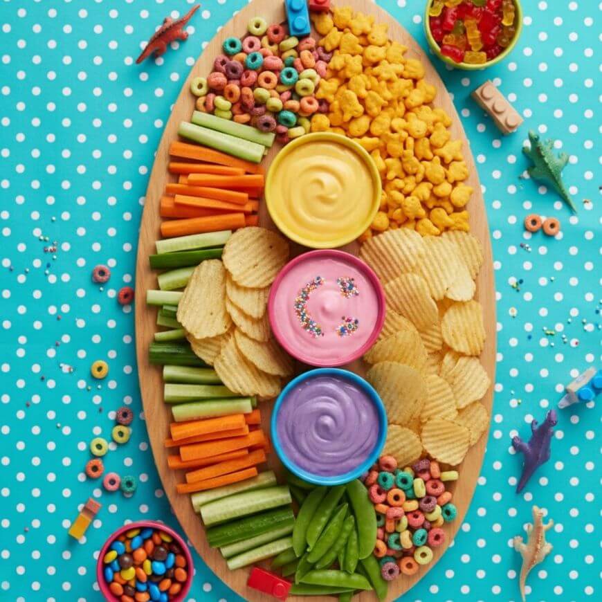 A colorful snack platter with potato chips, goldfish crackers, carrot and celery sticks, snap peas, three bowls of pastel dips with sprinkles, and fruit-flavored cereal, on a blue polka dot tablecloth with toy dinosaurs.