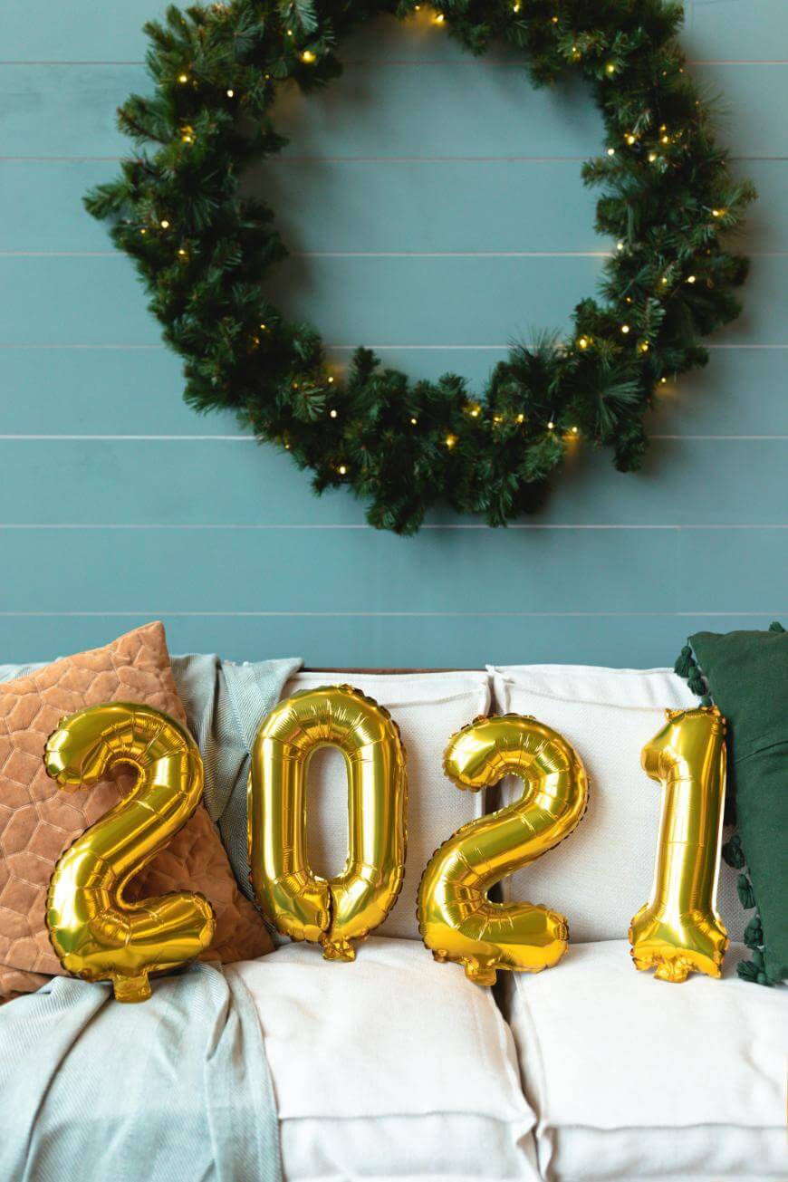 Gold balloon numbers spelling 2021 sit on a white couch with pillows. A large green wreath with lights hangs on the blue wall behind the couch.