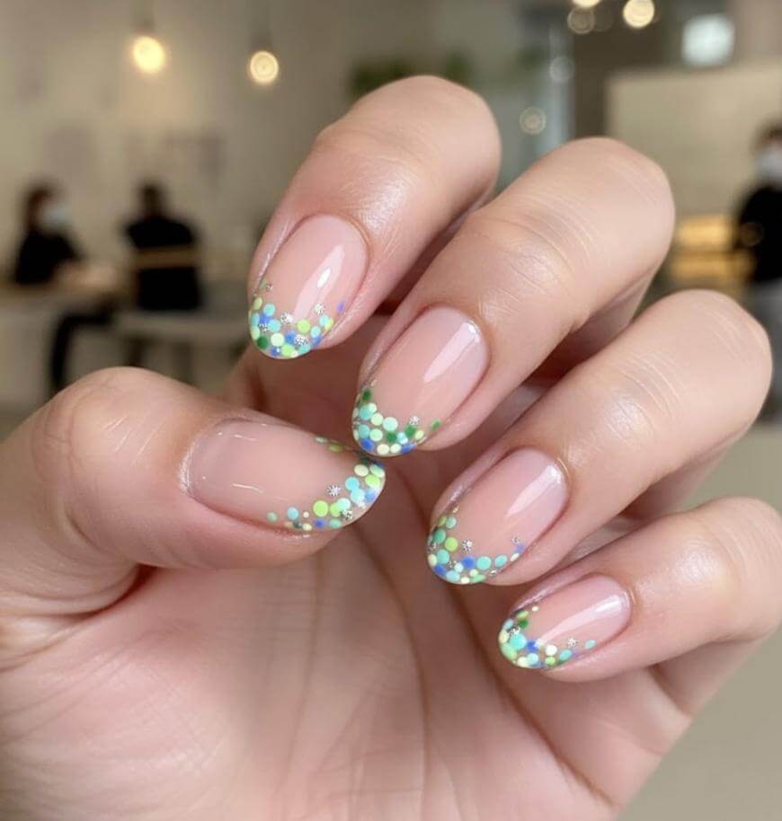 A hand with neatly manicured nails featuring a nude base and colorful, confetti-like green, blue, and yellow dots on the tips. The background is blurred, showing people sitting indoors.