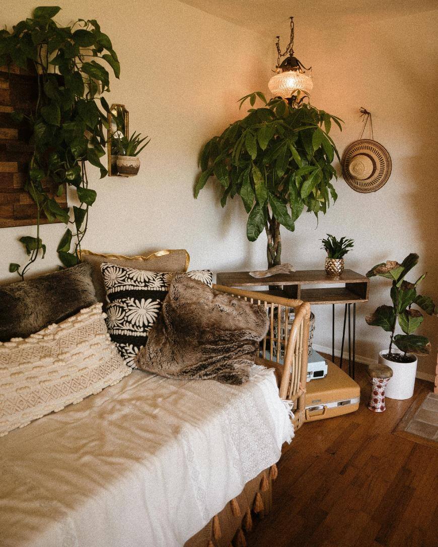 Cozy living room with a white daybed, textured pillows, wooden furniture, lush potted plants, a woven wall hat, a pineapple on a table, warm lighting, and wooden flooring.