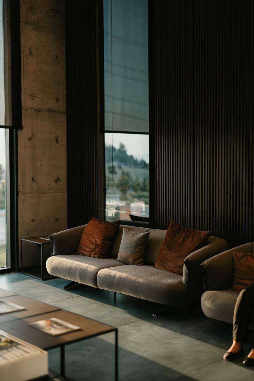 A modern, sunlit lounge features a gray sofa with brown pillows, set against dark vertical wall panels and tall windows. A coffee table with magazines sits in front, and a partial view of a person is visible on the right.