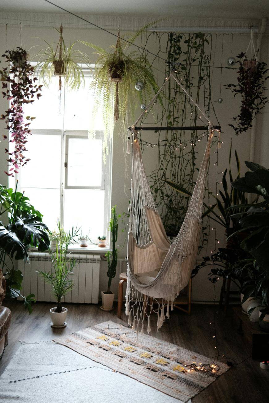A cozy room with a hanging macrame chair, surrounded by various potted plants and greenery. Natural light streams through a window, and string lights add a warm touch. A rug lies on the wooden floor beneath the chair.