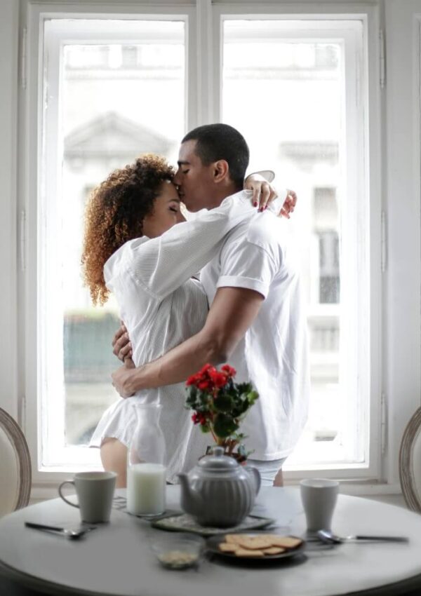 A couple stands by a window embracing and kissing, both dressed in white shirts. In the foreground, a table is set with a teapot, cups, a red flower, milk, and plates of food.