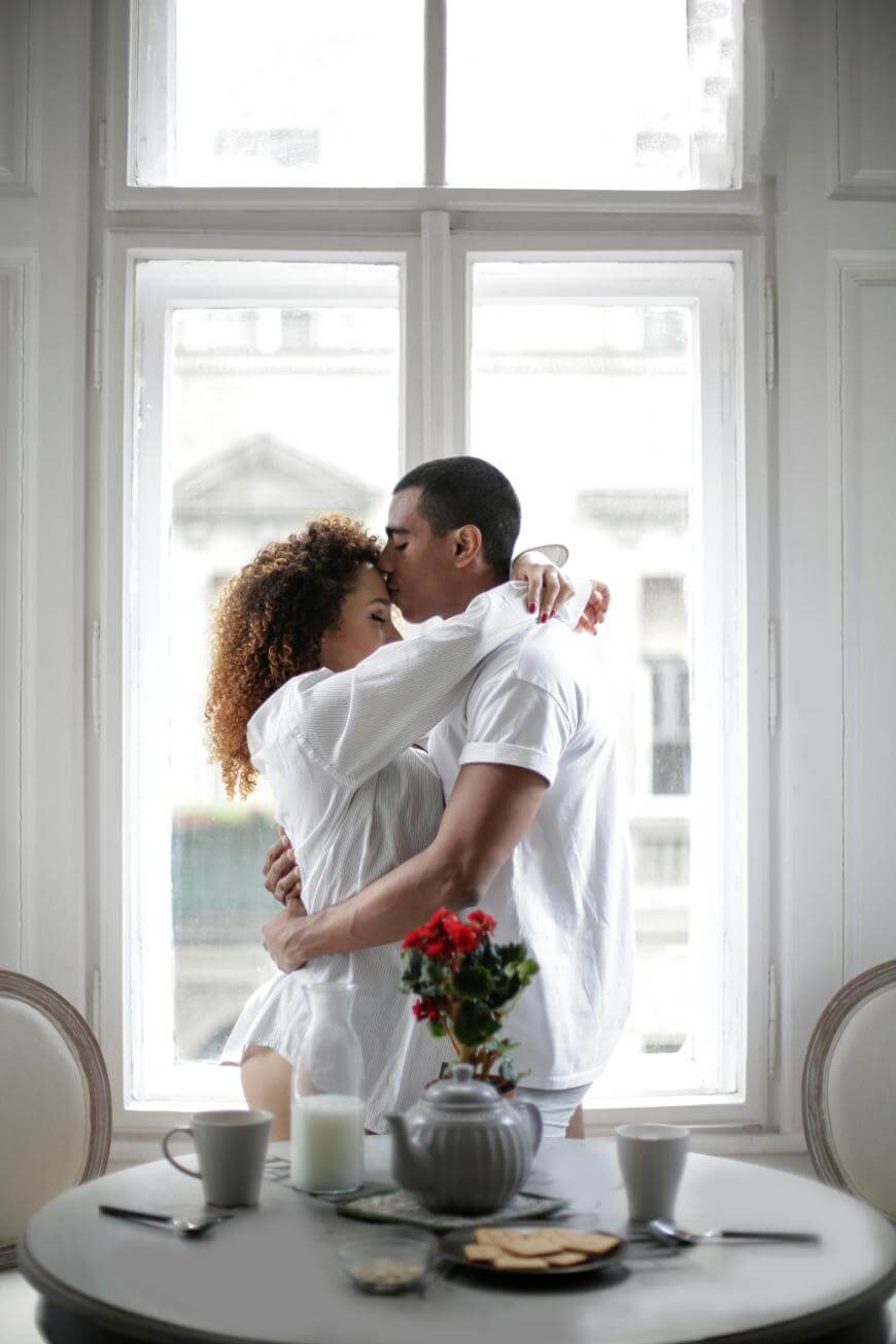 A couple stands by a large window, embracing and kissing. They are wearing white shirts, with a breakfast table set with a teapot, cups, and a vase of red flowers in the foreground.