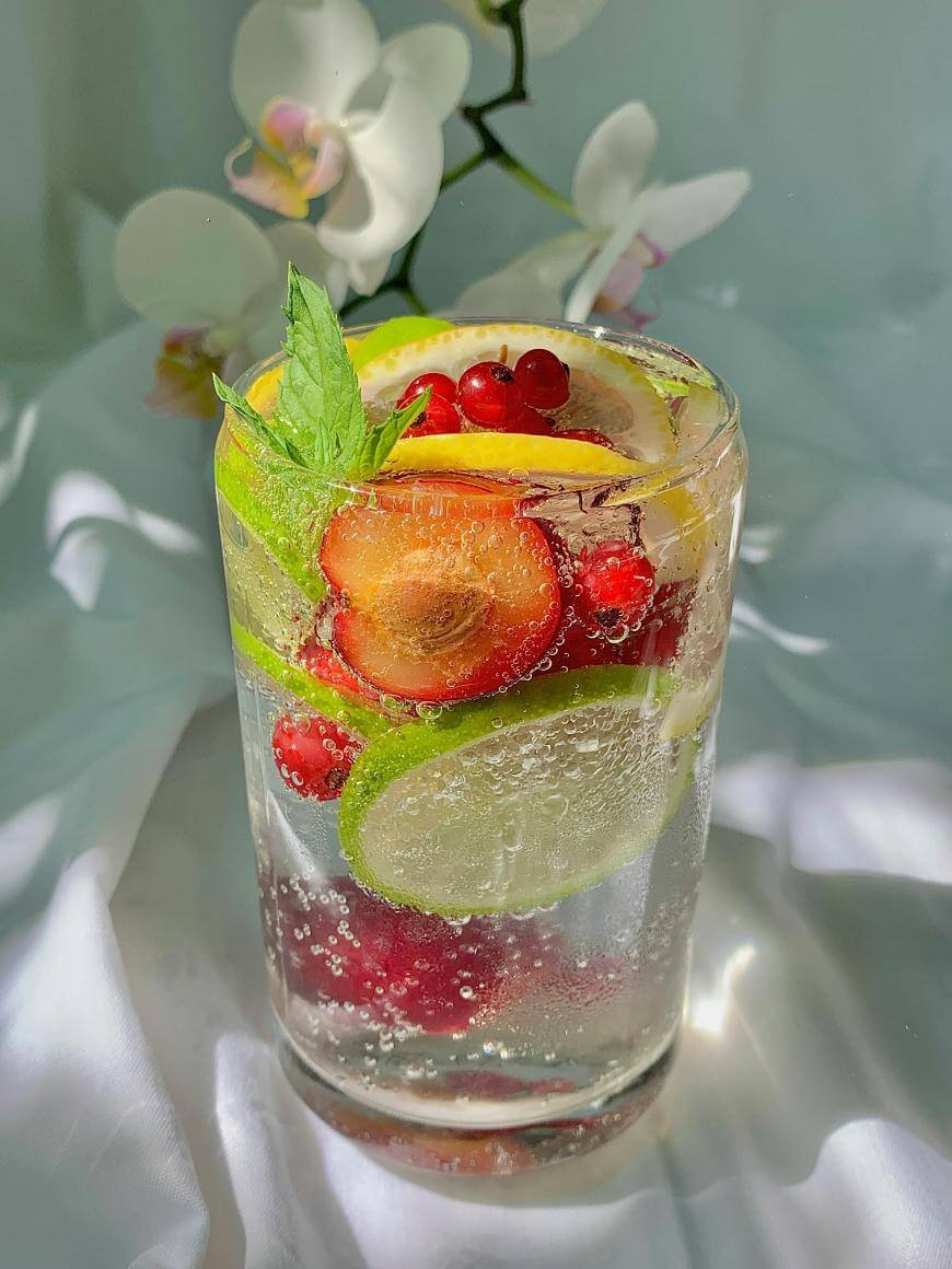 A glass of sparkling water filled with slices of lime, lemon, and strawberry, along with red currants and a sprig of mint. White orchid flowers are blurred in the background. Light creates soft reflections on the surface.