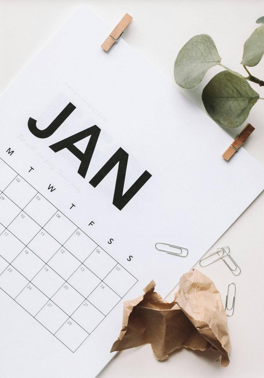 A minimalistic calendar open to January lies on a white surface, decorated with a green leafy branch, paper clips, and a crumpled piece of brown paper.