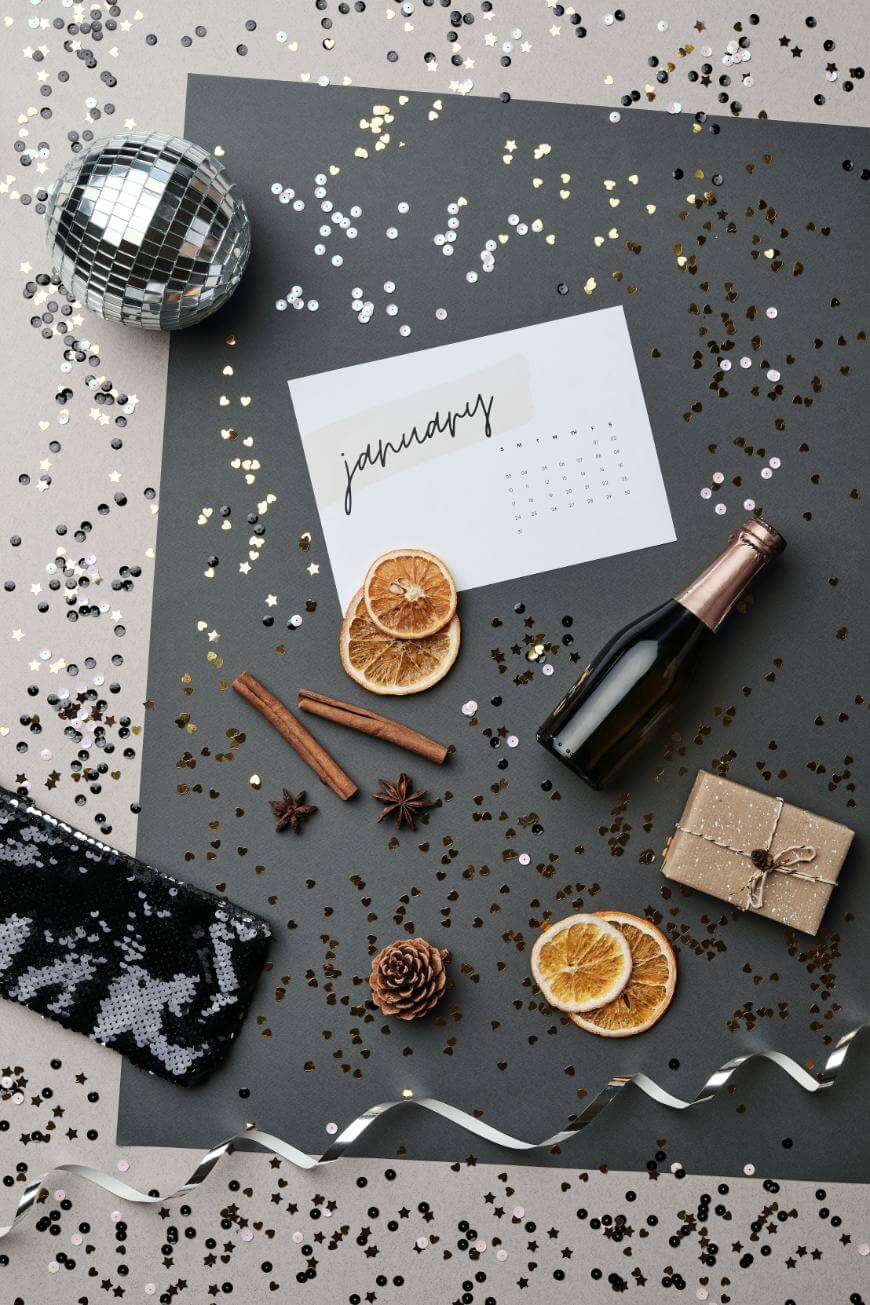 Flat lay of New Year’s celebration items: a January calendar, mini champagne bottle, dried orange slices, cinnamon sticks, star anise, disco ball, sequined purse, gold gift box, pine cone, ribbon, and confetti on gray paper.
