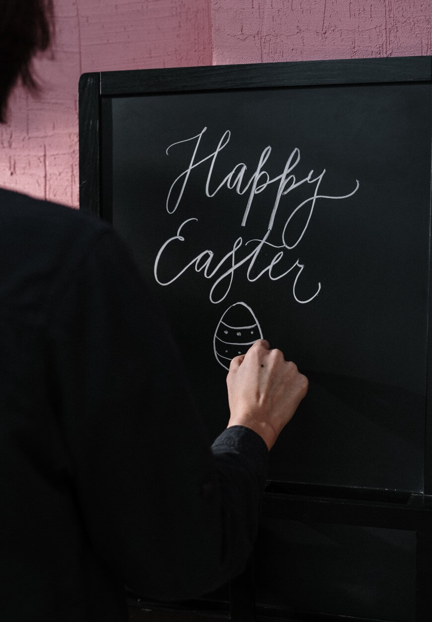 A person writes Happy Easter in cursive on a black chalkboard, adding a simple drawing of a decorated Easter egg underneath the text.