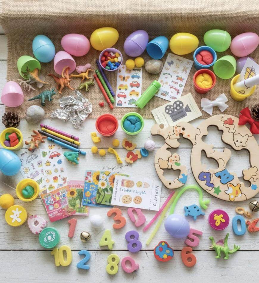 A colorful assortment of children’s toys and activities, including plastic eggs, wooden puzzles, playdough, animal figurines, pom-poms, numbers, stickers, crayons, and small books spread out on a light surface.