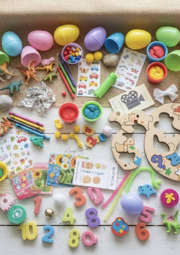 A variety of colorful toys and crafts, including Easter eggs, dinosaur figurines, numbers, stickers, playdough, colored pencils, and puzzle pieces, are spread out on a white wooden surface.