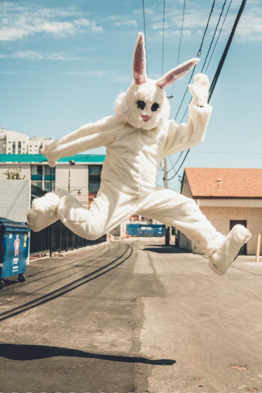 A person in a white bunny costume is jumping high in the air in the middle of an empty urban alleyway on a sunny day.