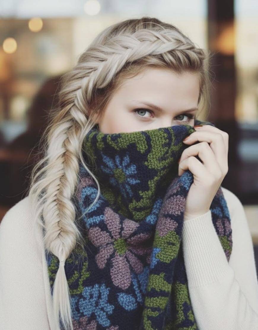 A woman with long blonde hair in a fishtail braid partially covers her face with a blue, green, and purple floral-patterned scarf, leaving only her eyes visible.