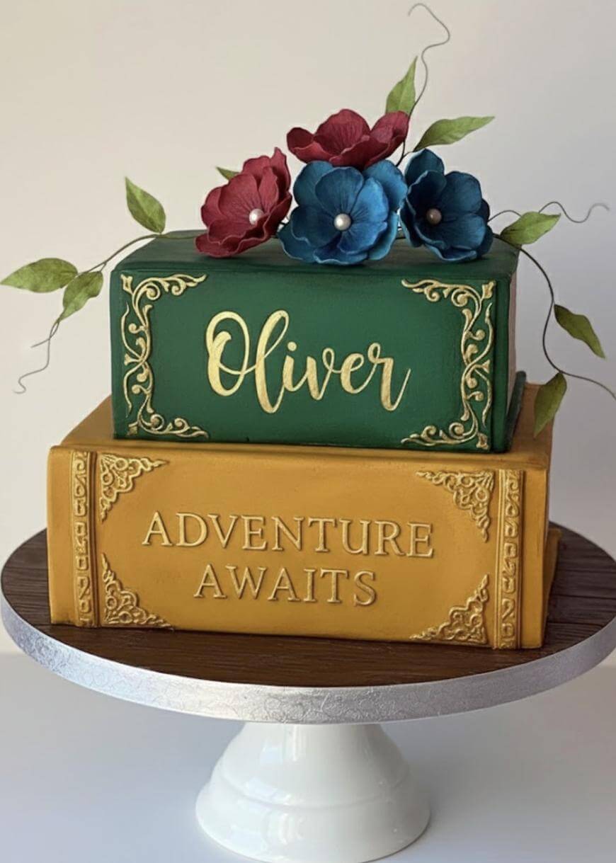 A cake designed to look like two stacked books, the top green book with Oliver written in gold and the bottom yellow book with Adventure Awaits. Red and blue sugar flowers and green leaves decorate the top.