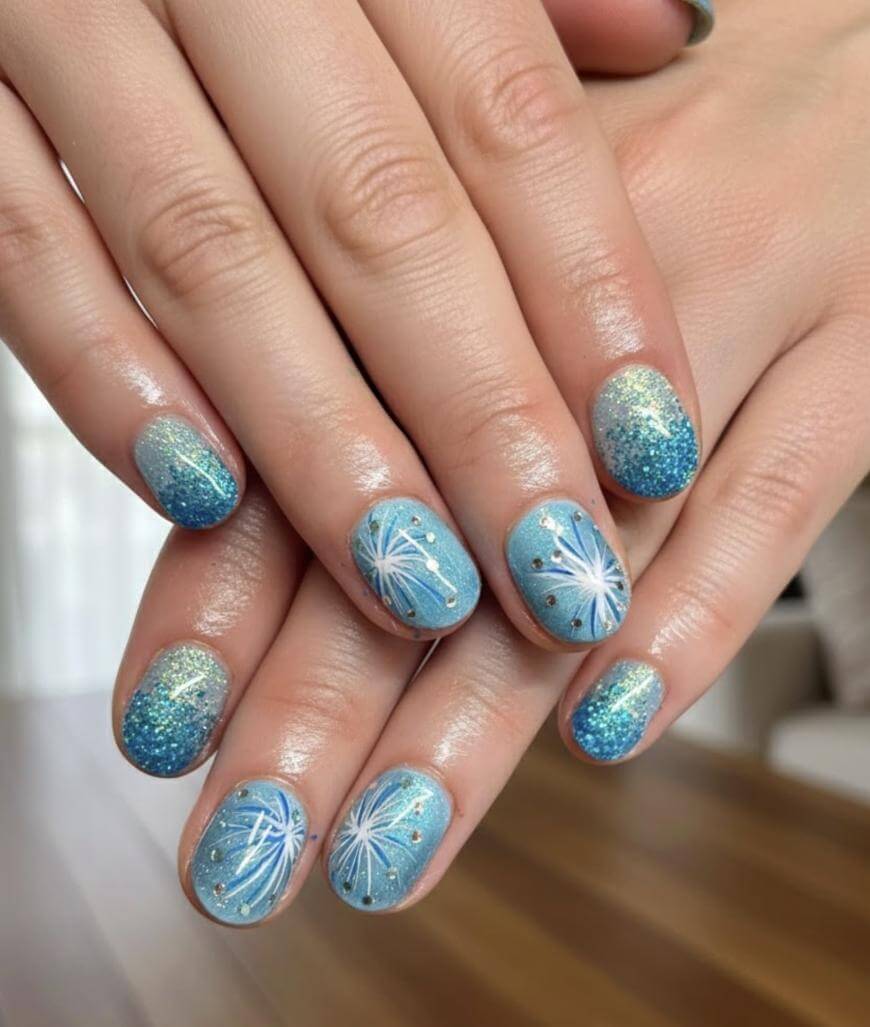 Close-up of hands with short, rounded nails painted blue and glittery, featuring white floral and dandelion-like designs on some nails. The nails have a glossy finish and look freshly manicured.