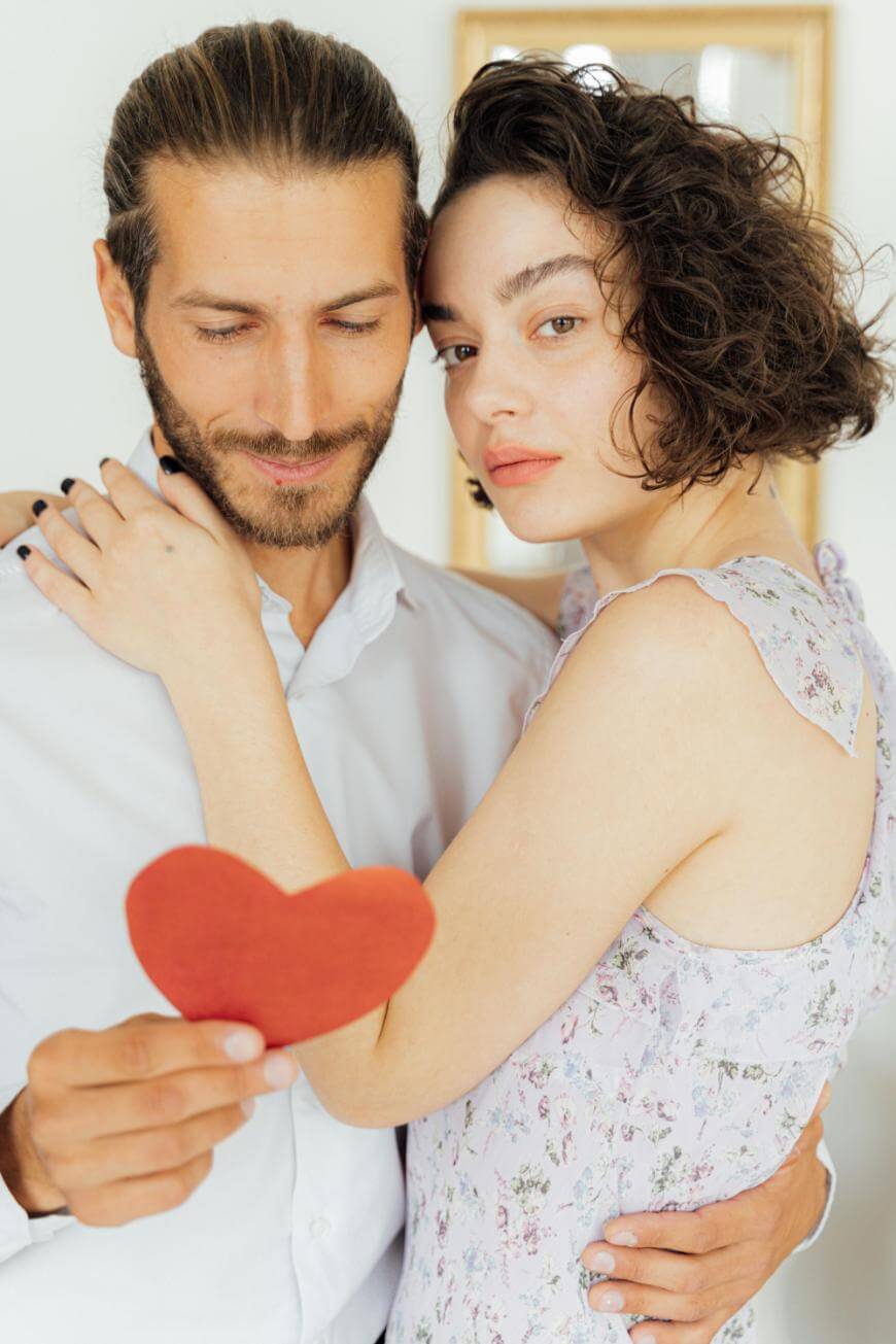 A man and a woman stand close together, embracing. The woman has one arm around the mans neck and looks at the camera, while the man looks down at a red paper heart he is holding.