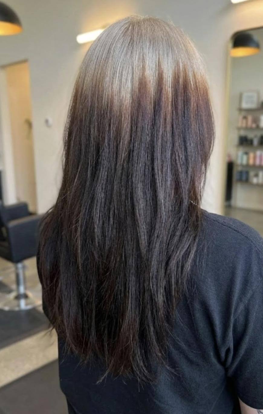 A person with long, straight hair featuring a blend of gray at the roots, transitioning to brown and then to dark brown at the ends, is seen from behind in a modern salon setting.