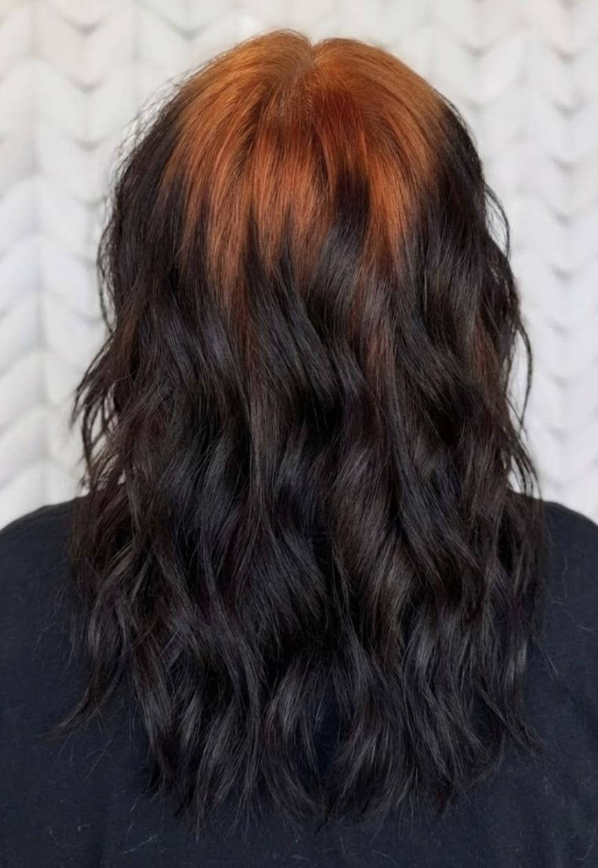 A person with wavy, shoulder-length hair dyed black with a distinct copper-orange section at the roots and top. The photo is taken from the back against a light, patterned background.