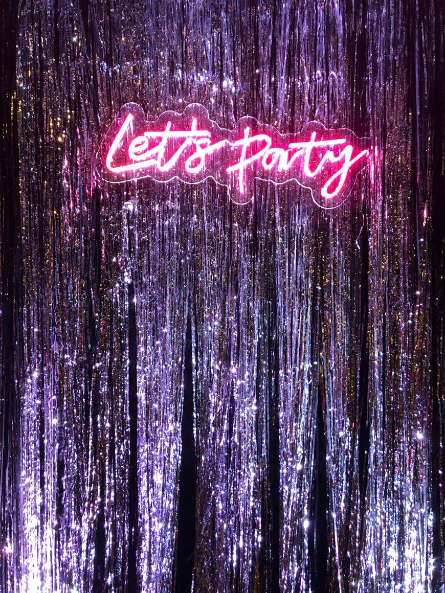 A neon pink sign reading Lets Party glows above shiny silver tinsel streamers, creating a festive party backdrop.