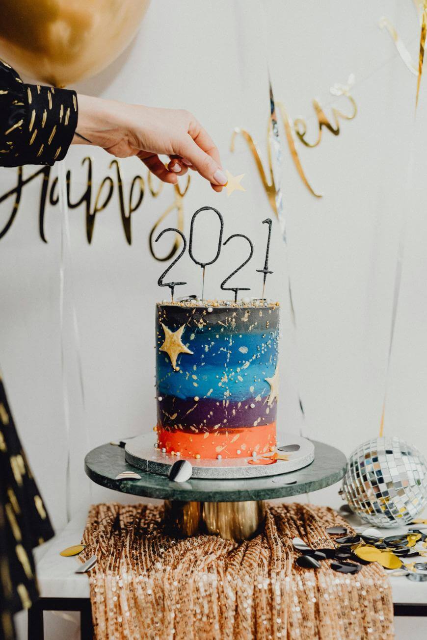 A hand lights candles shaped as 2021 on a colorful, star-decorated cake. The cake sits on a table with gold decor, a disco ball, and Happy New Year balloons in the background.