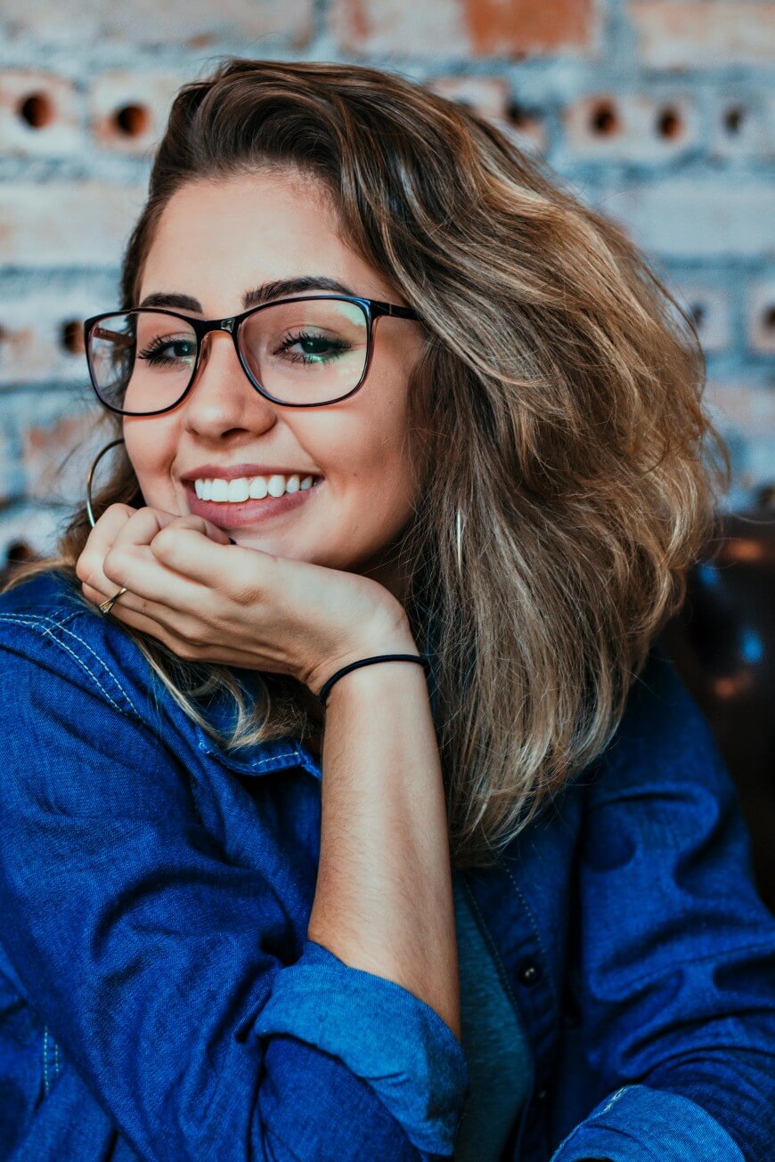 A young woman with wavy light brown hair and glasses smiles warmly, resting her chin on her hand. She wears a blue denim shirt and sits in front of a brick wall background.