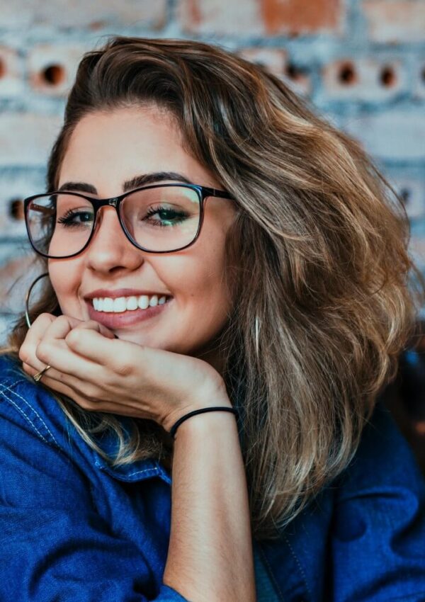 A young woman with wavy light brown hair and glasses smiles, resting her chin on her hand. She is wearing a blue denim shirt and is sitting in front of a brick wall.