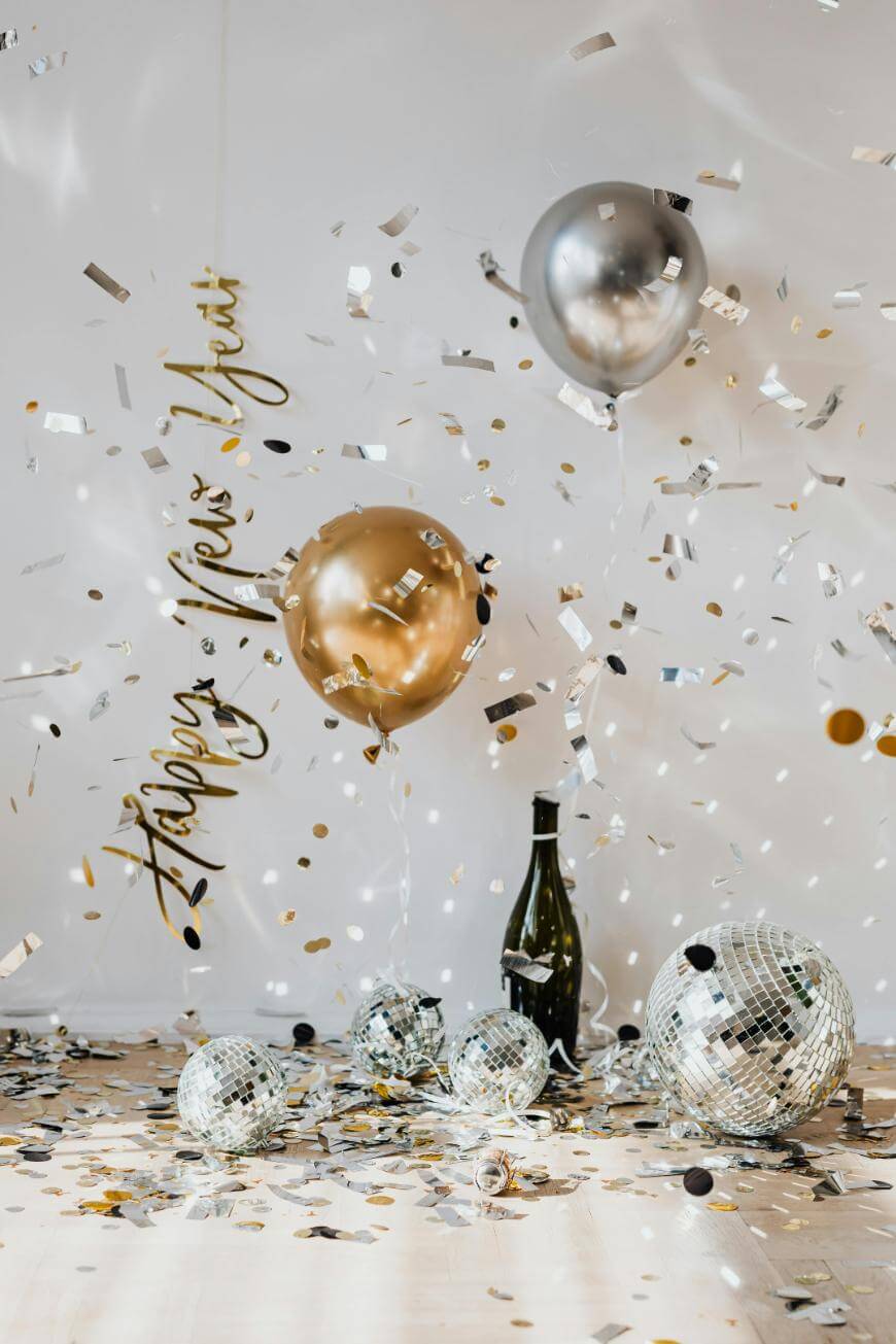 Gold and silver balloons, disco balls, confetti, and a champagne bottle create a festive New Year’s celebration scene. A shiny Happy New Year sign hangs on the wall in the background.